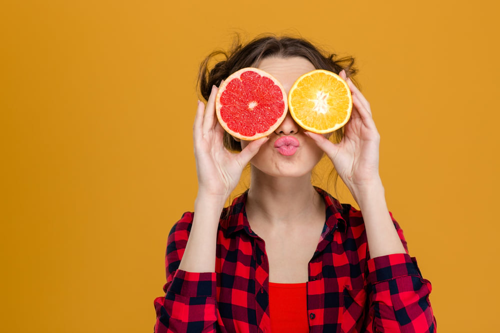 Funny playful young woman in checkered shirt holding halves of citrus fruits against her eyes and making duck face over yellow background-1